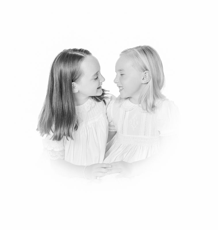 Two young girls in white dresses share a sweet moment, smiling at each other in a soft black and white portrait.