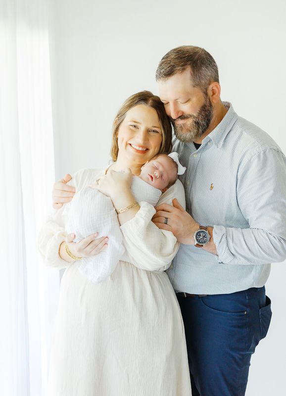 A smiling couple in a bright studio tenderly holds their sleeping newborn baby, dressed in white.