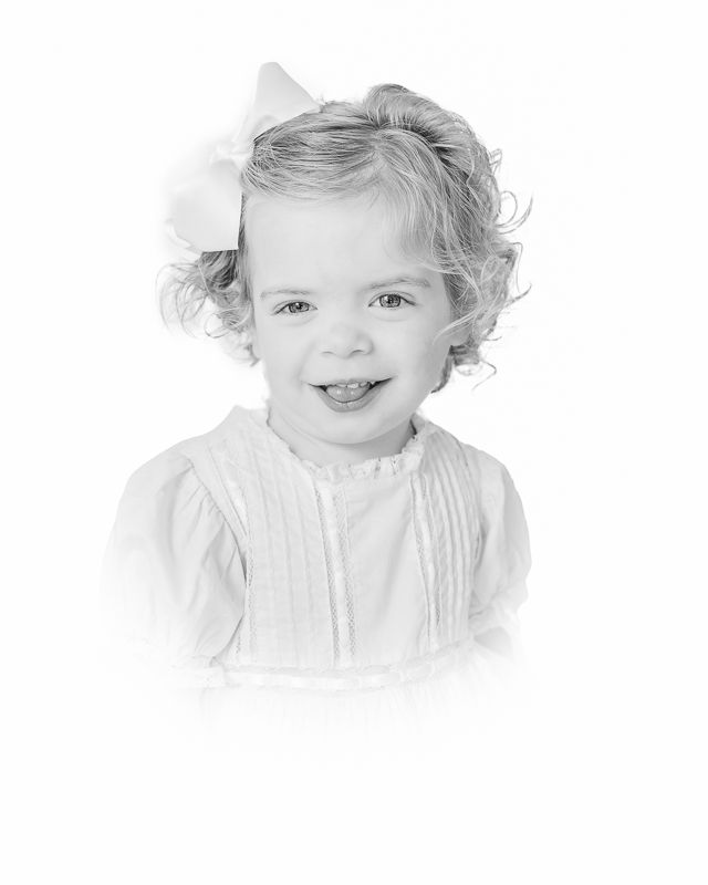 Black and white portrait of a smiling toddler girl with curly hair and a white bow, wearing a white dress.