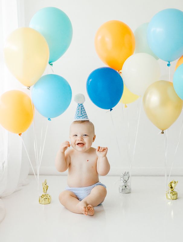 Smiling baby in birthday hat sits surrounded by colorful blue, yellow, and gold balloons during first birthday cake smash.