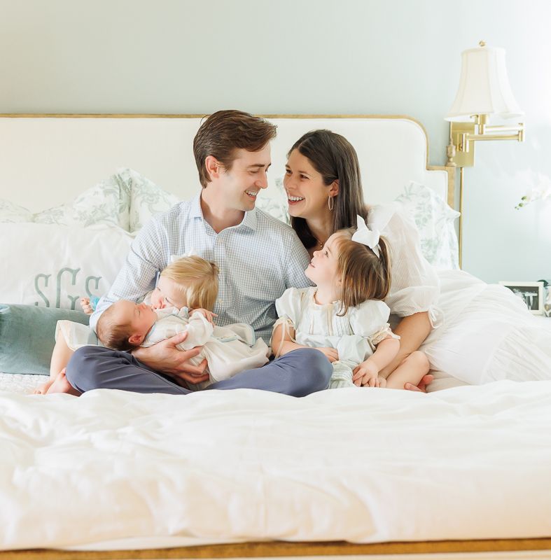 A happy family of four sits on a white bed, parents smiling at each other while holding their two young children.