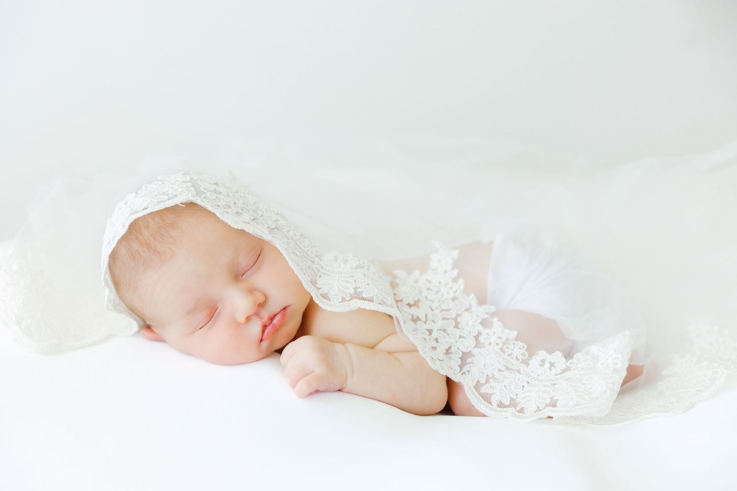 Sleeping newborn baby in white lace bonnet and outfit on soft white blanket.