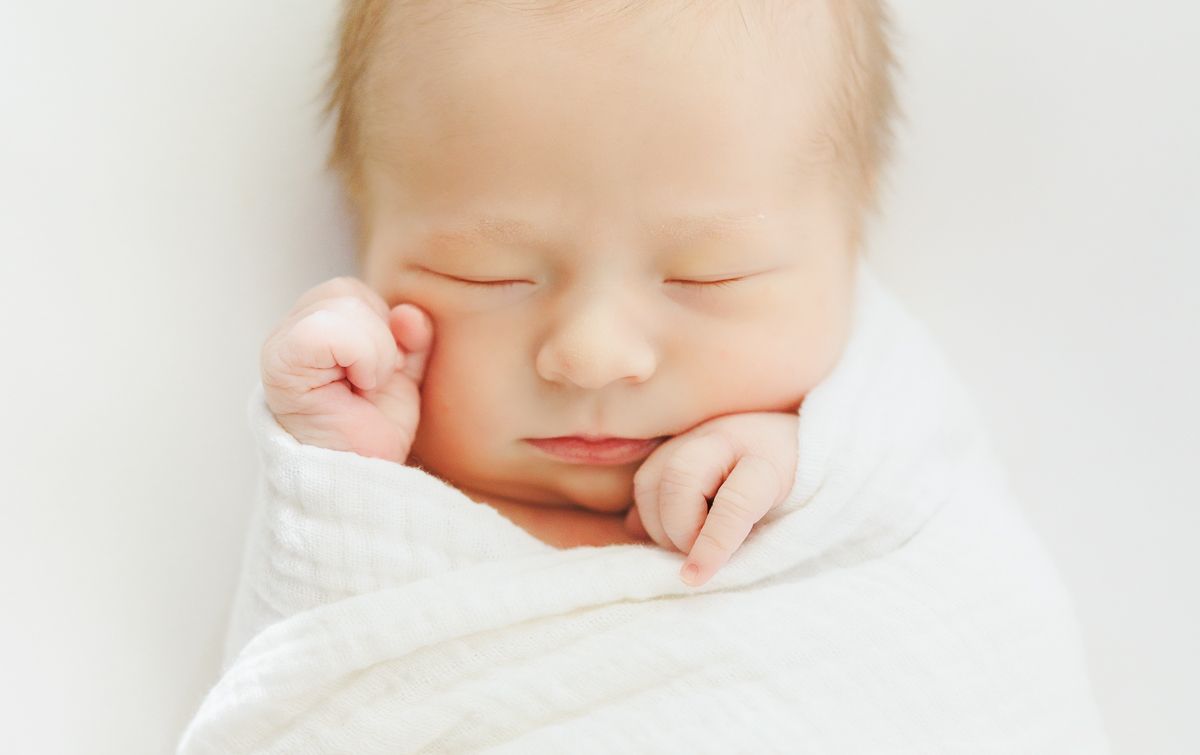 Peaceful newborn baby sleeping soundly, swaddled in a soft white blanket with tiny hands near face.