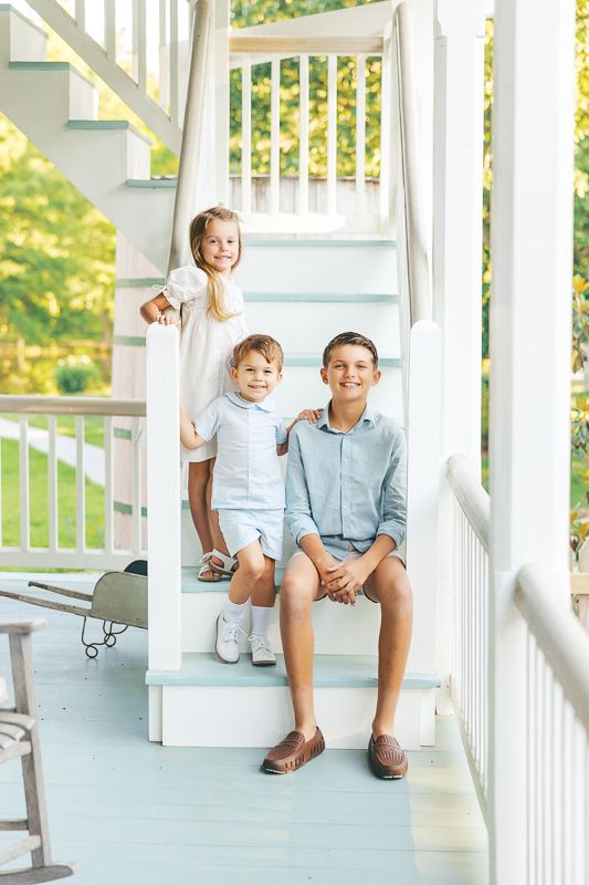 Three children pose on white porch steps, dressed in light blue and white outfits, surrounded by lush green trees.
