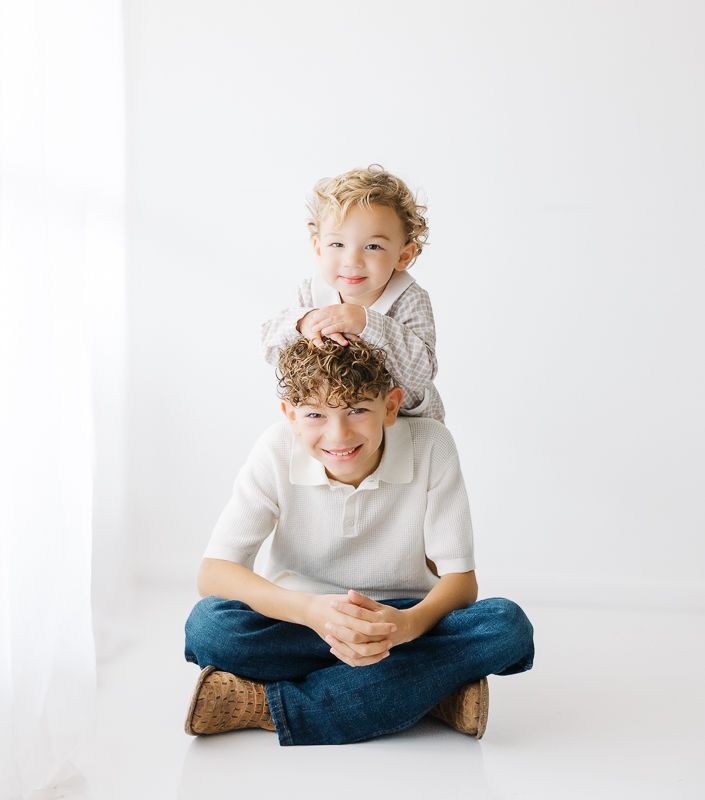 Two smiling brothers pose together, older boy sitting cross-legged with younger sibling resting on his shoulders.