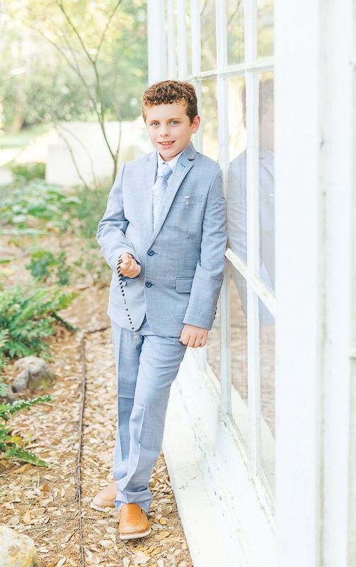 Boy in light blue suit and tan shoes leaning against white building outdoors for formal portrait photo.