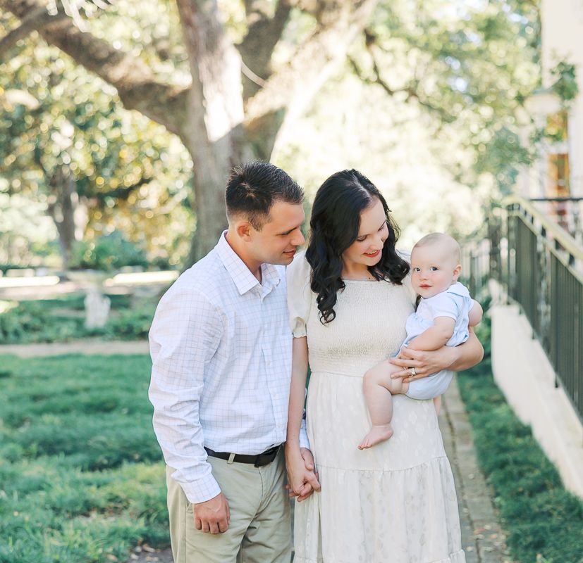 Young couple with baby exploring a sunlit garden path lined with hedges and large oak trees.