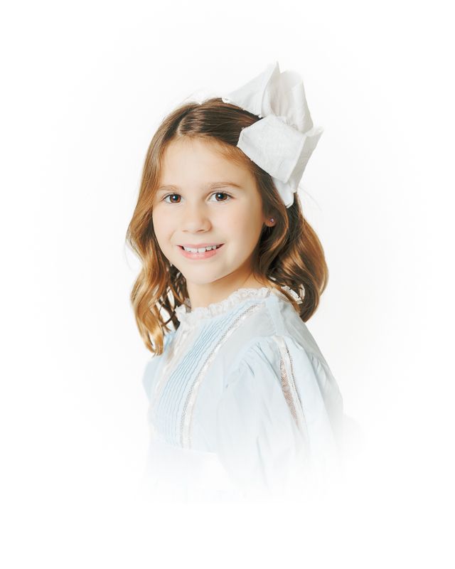Young girl smiling in a light blue dress with a white bow in her brown hair.