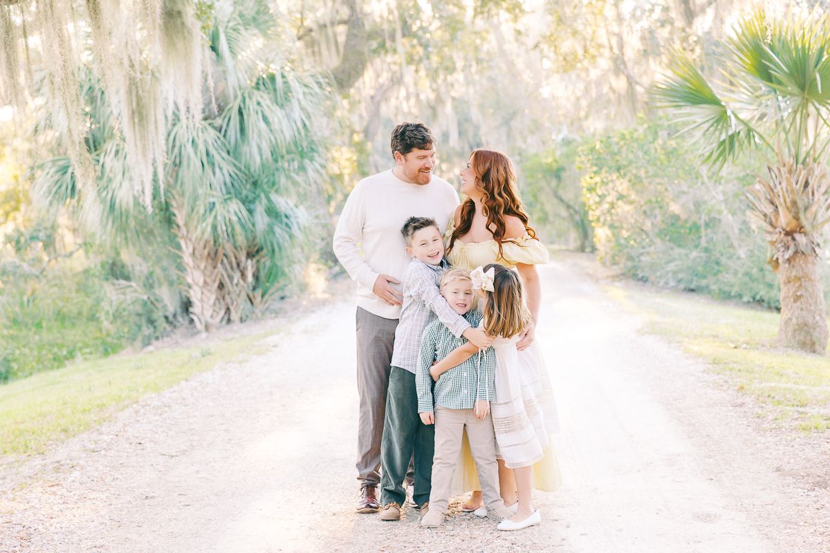 Family of four poses on a sunlit path surrounded by palm trees and Spanish moss in a bright outdoor portrait.