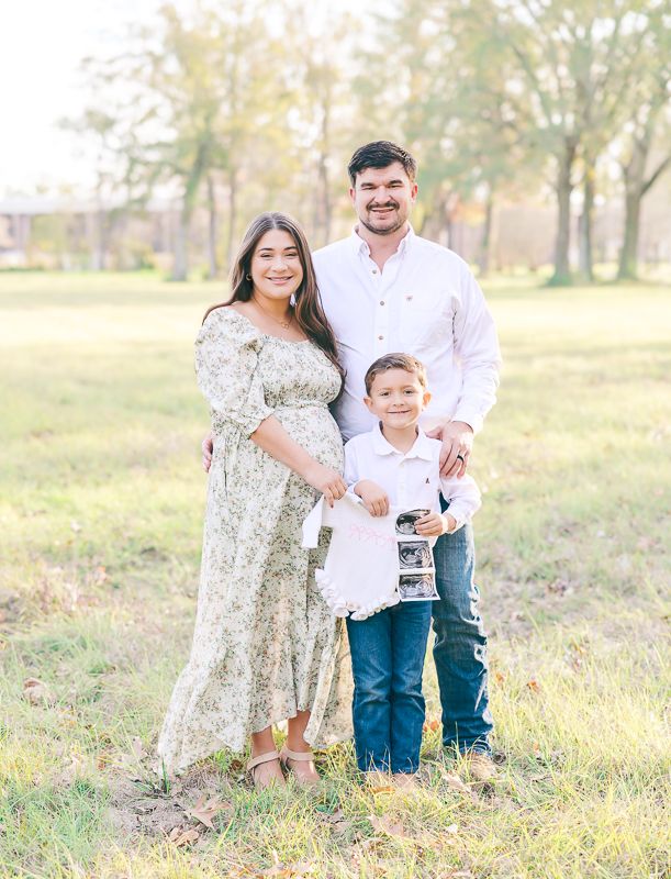 Expecting family of three posing outdoors holding ultrasound photos in a sunny open field setting.