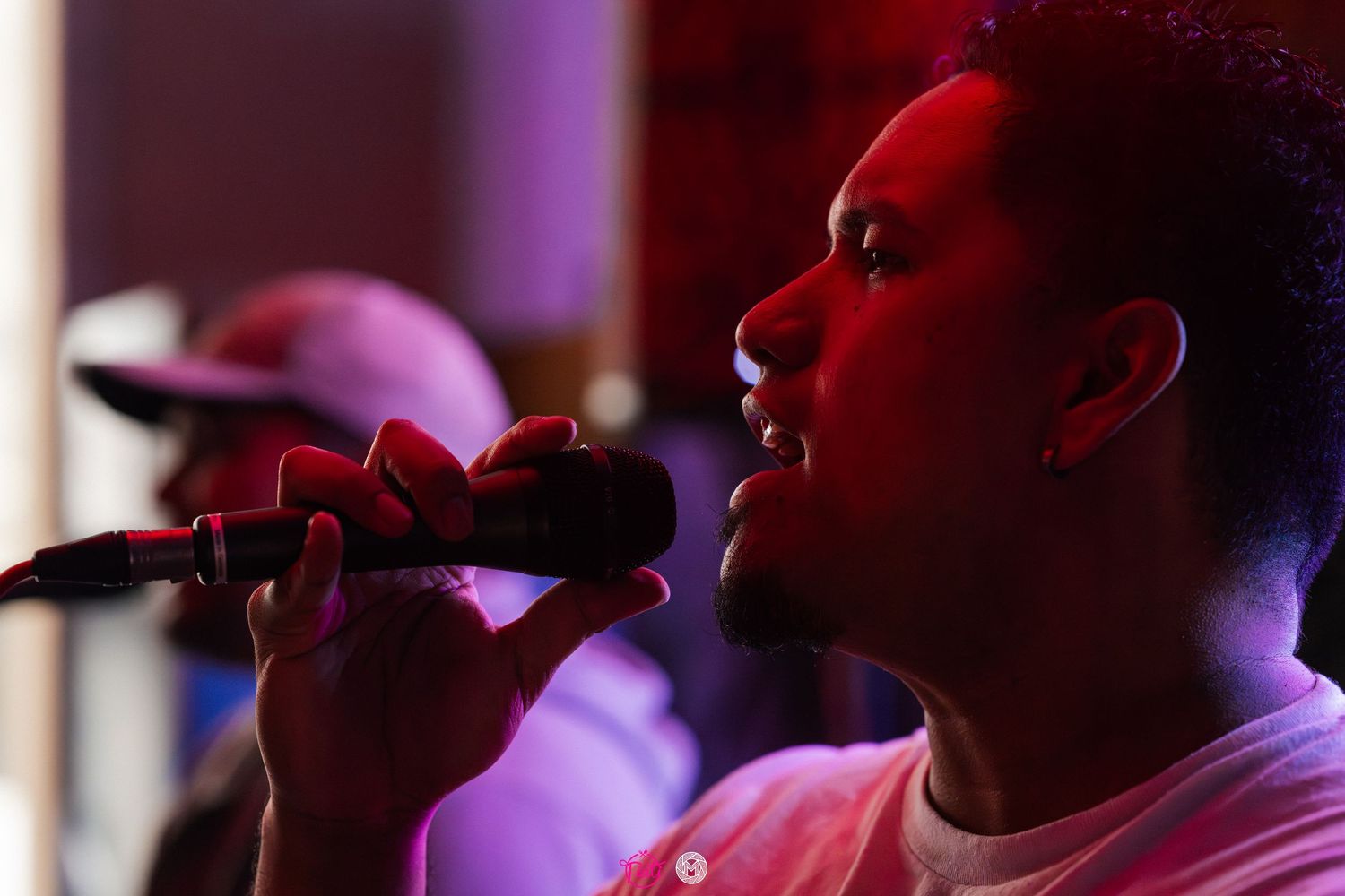 Young man singing passionately into a microphone under red stage lighting at a live music event.