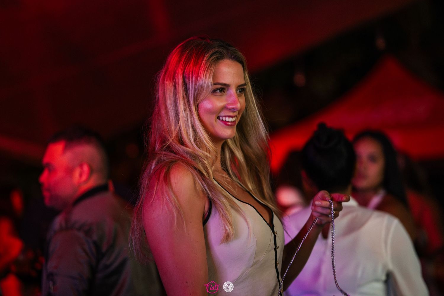 Blonde woman smiling and holding a bag under red ambient lighting at a night event.