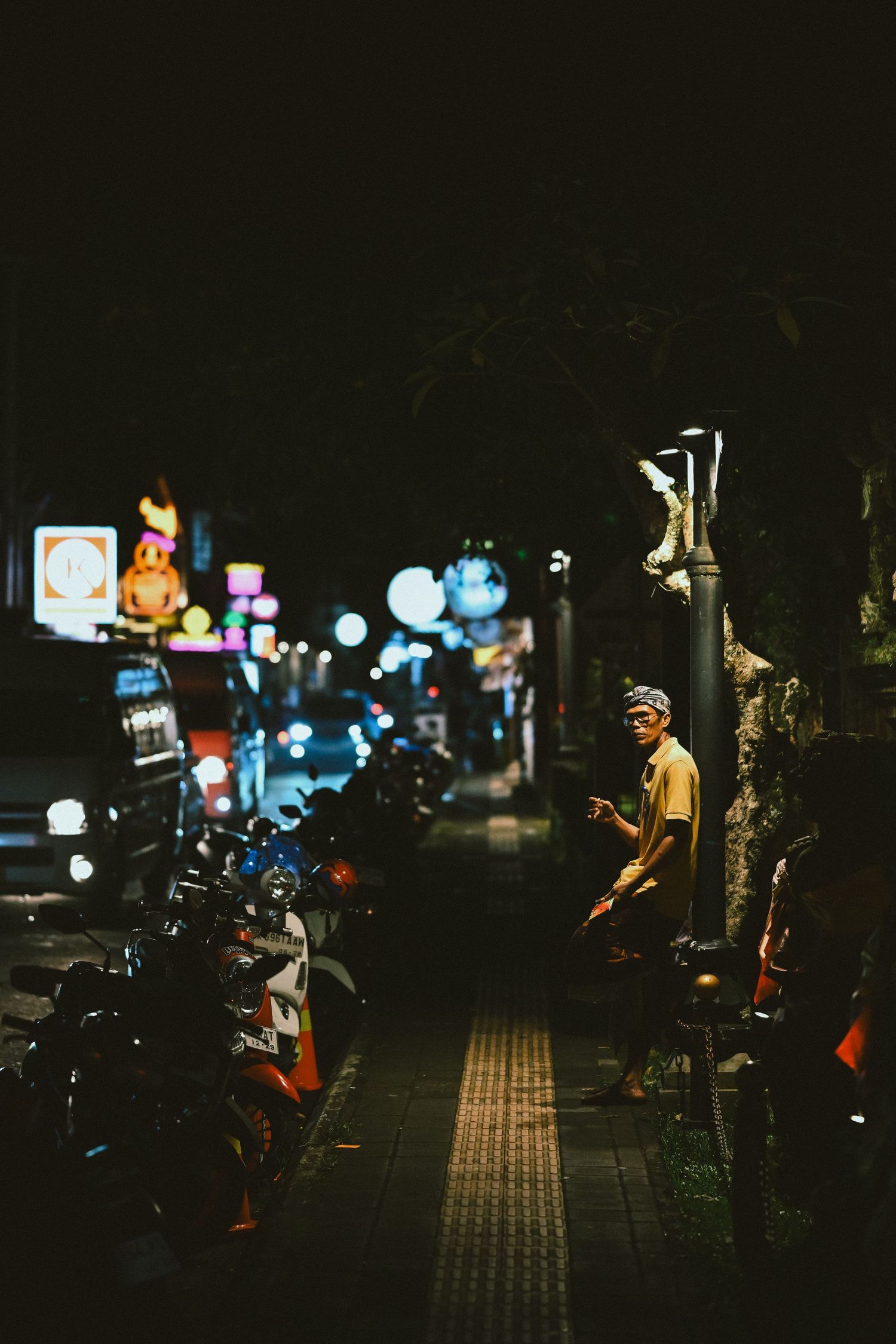 Man sitting under street lamp on busy Asian city street at night with motorcycles.