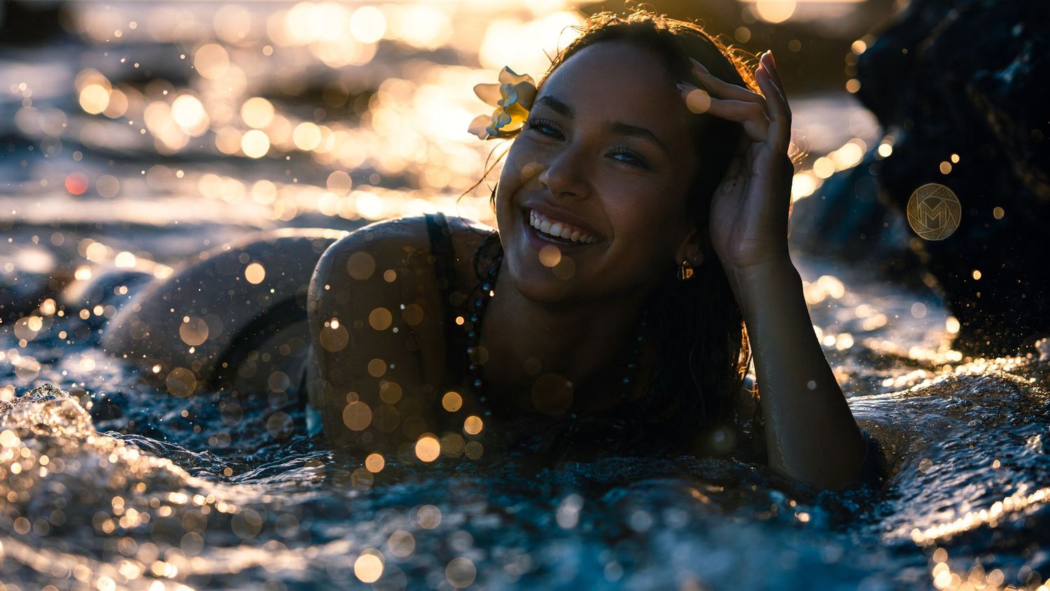 Smiling woman with flower in hair lying in sparkling shallow ocean water at sunset.