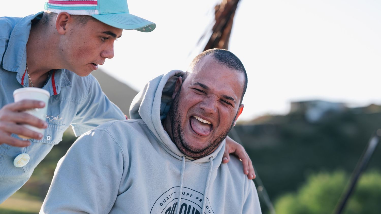 Two men laughing together outdoors with scenic green hills in the background.
