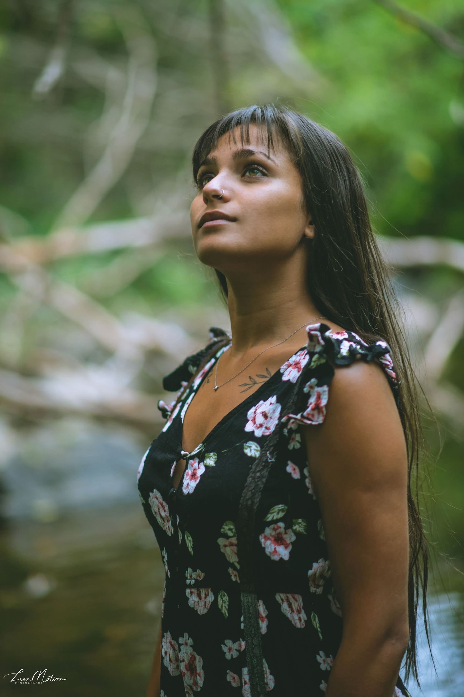Young woman in floral dress gazing upward in a lush green forest by a stream.
