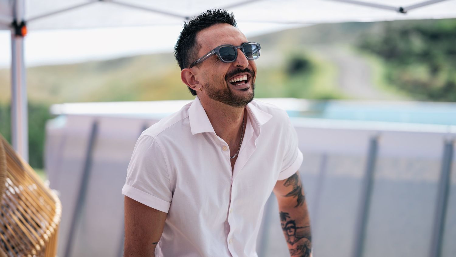 Smiling tattooed man in white shirt and sunglasses laughing outdoors at a sunny social event.
