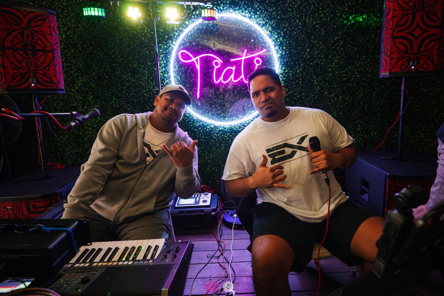 Two men posing together in front of glowing Tiato neon sign with keyboard setup.