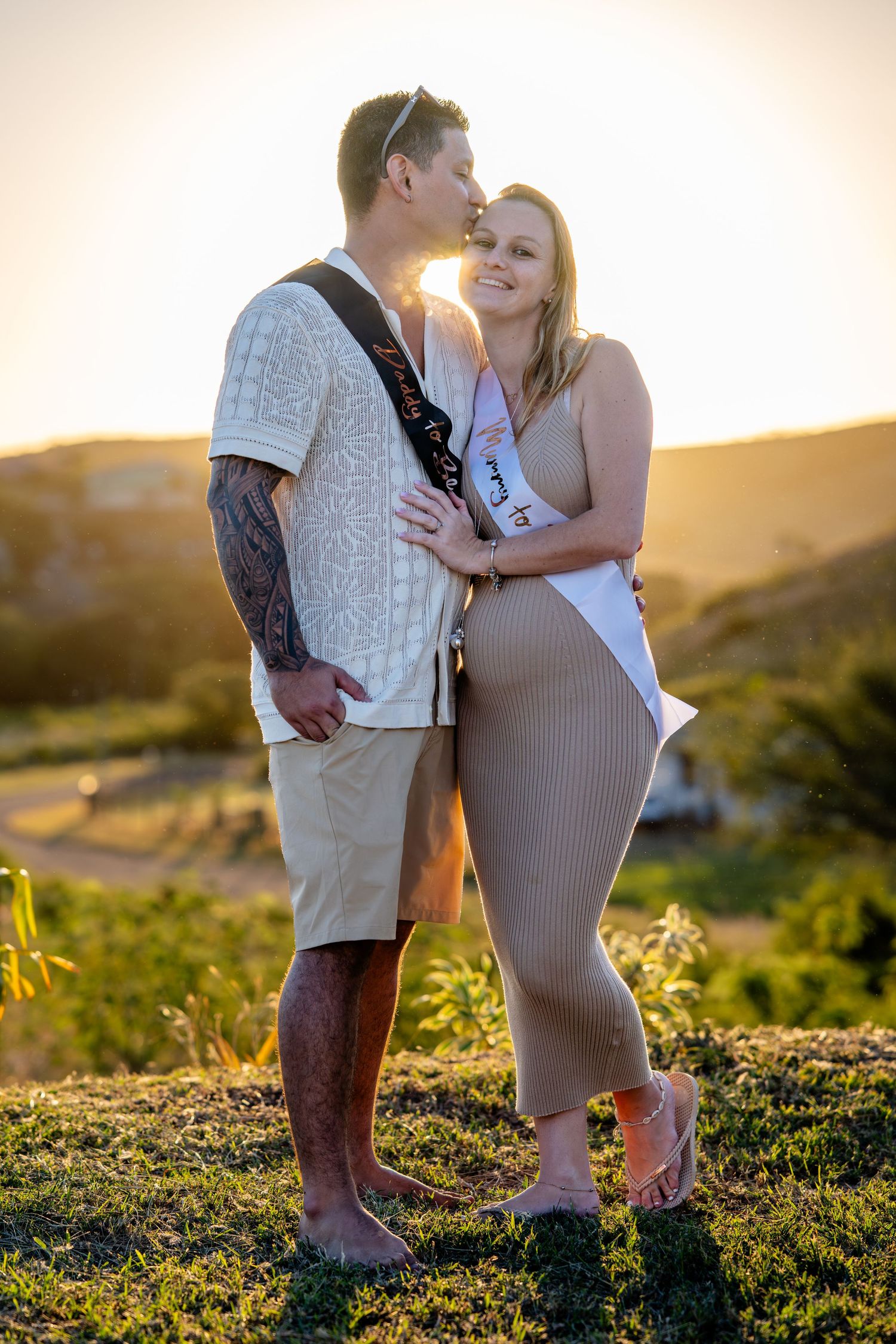 Couple embracing at golden sunset with woman wearing bride-to-be sash in scenic outdoor setting.