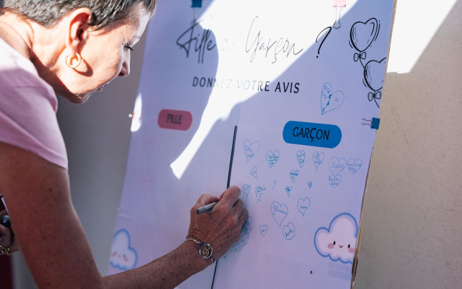 Woman writing on gender reveal board with fille or garcon text at outdoor party.