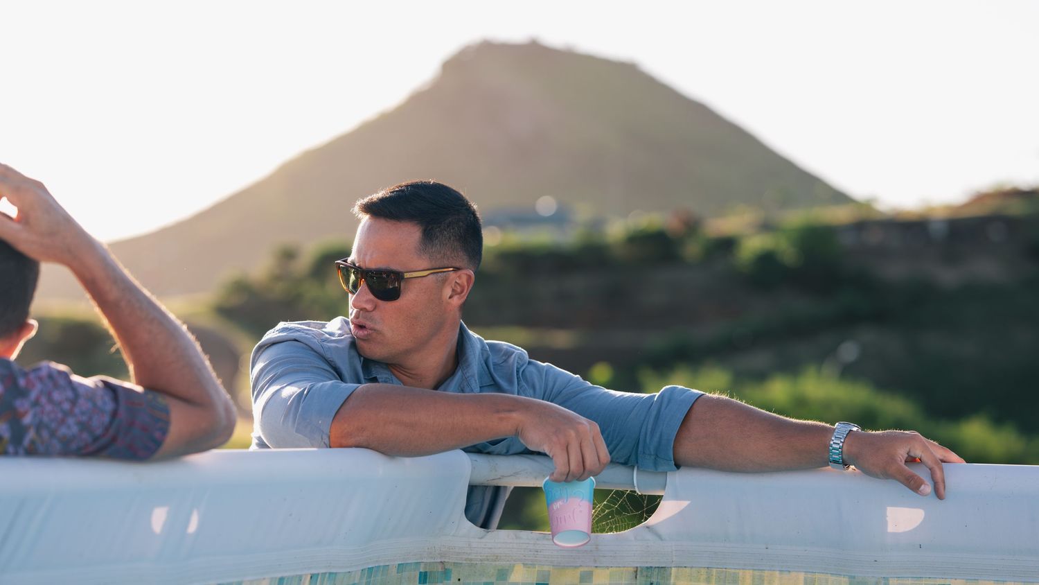 Man in sunglasses and grey shirt leaning on white railing with mountain sunset backdrop.