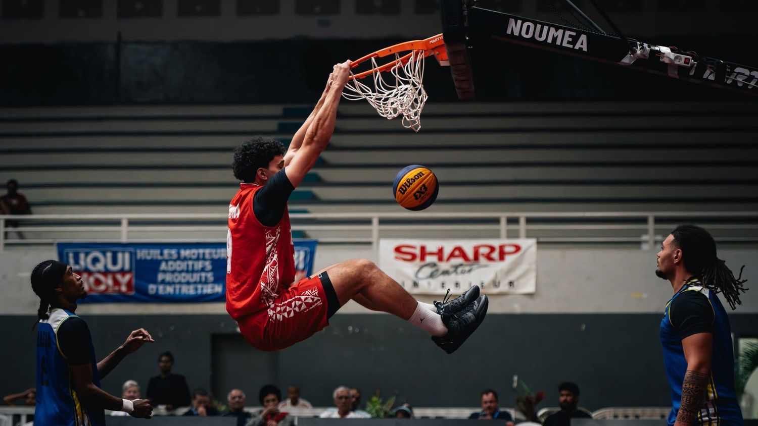 Basketball player performing powerful slam dunk during indoor game in Noumea arena.