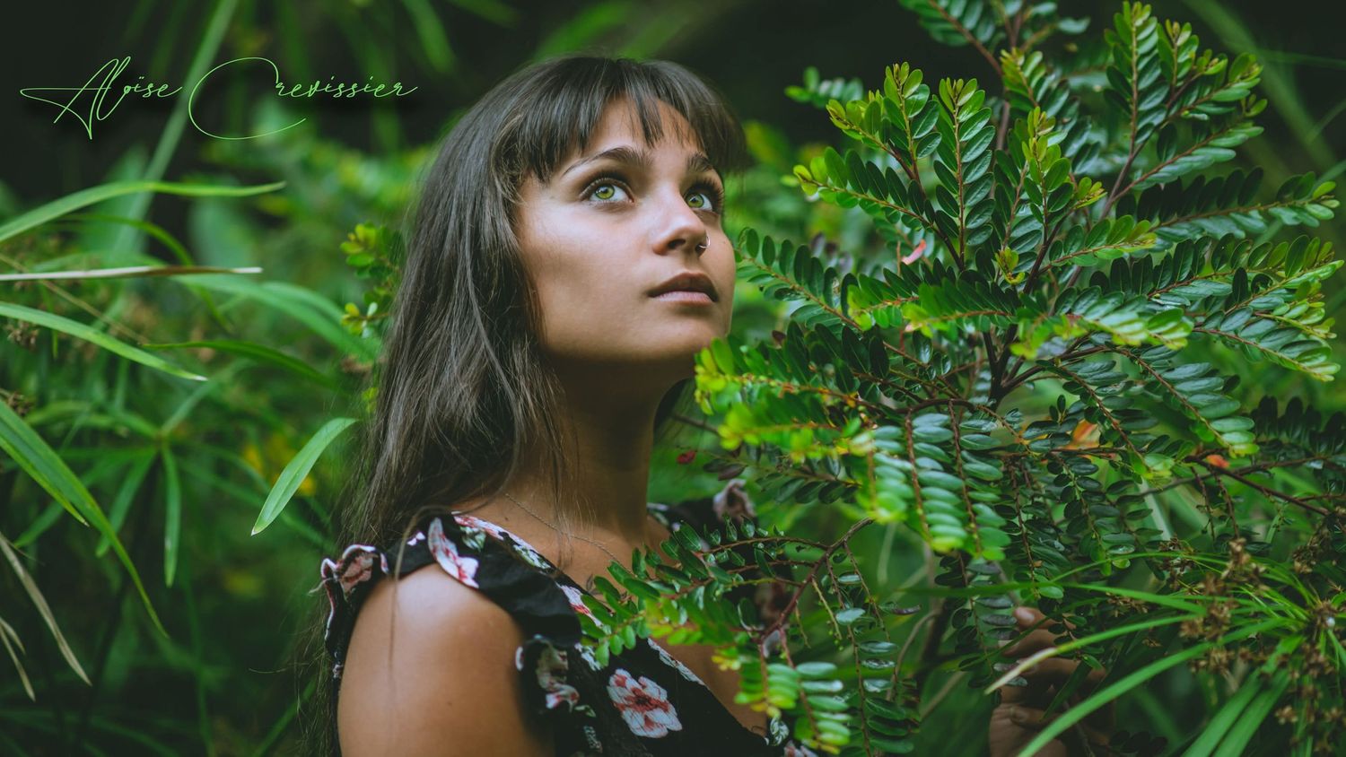 Young woman in floral dress surrounded by vibrant green tropical foliage looking upward.