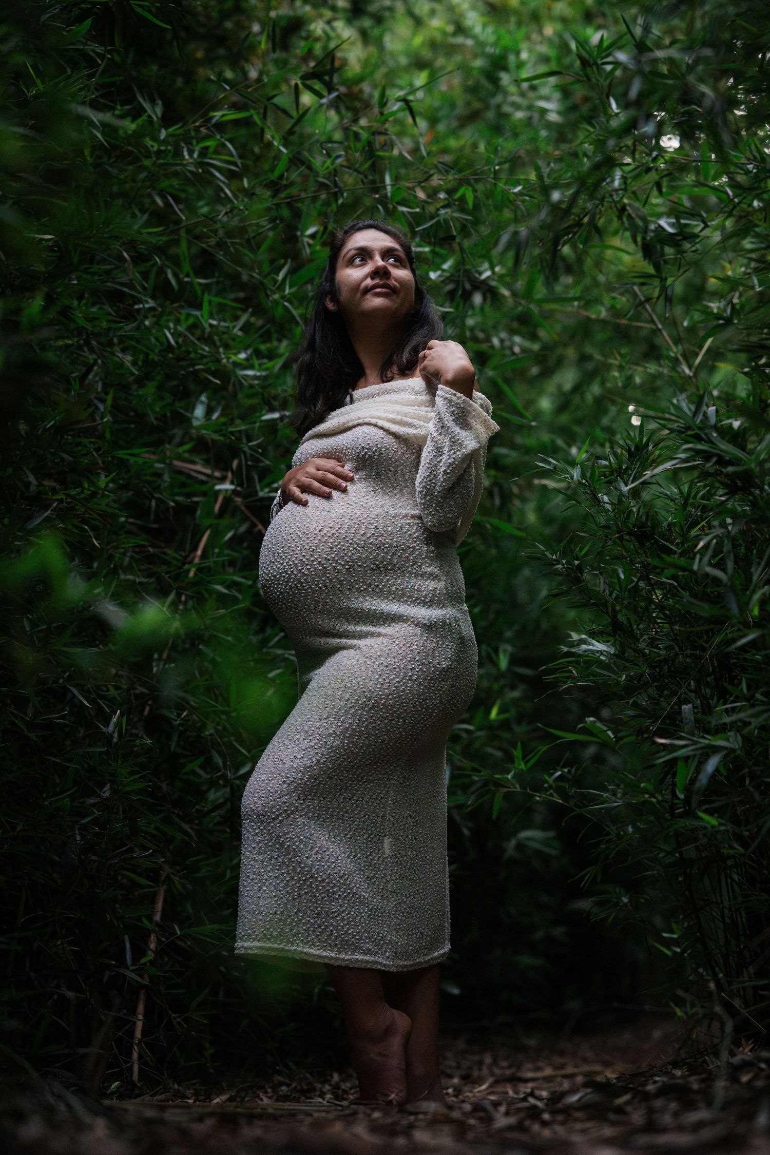 Pregnant woman in white knit dress standing barefoot among lush green bamboo foliage.