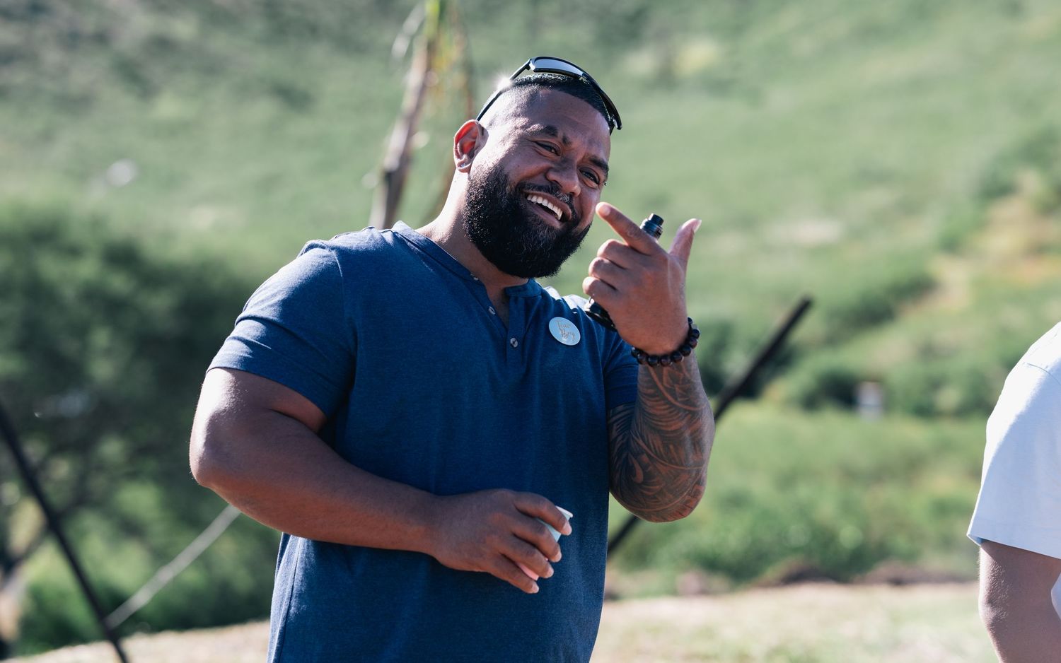 Smiling man in navy blue polo shirt giving shaka hand gesture outdoors with green hills in background.