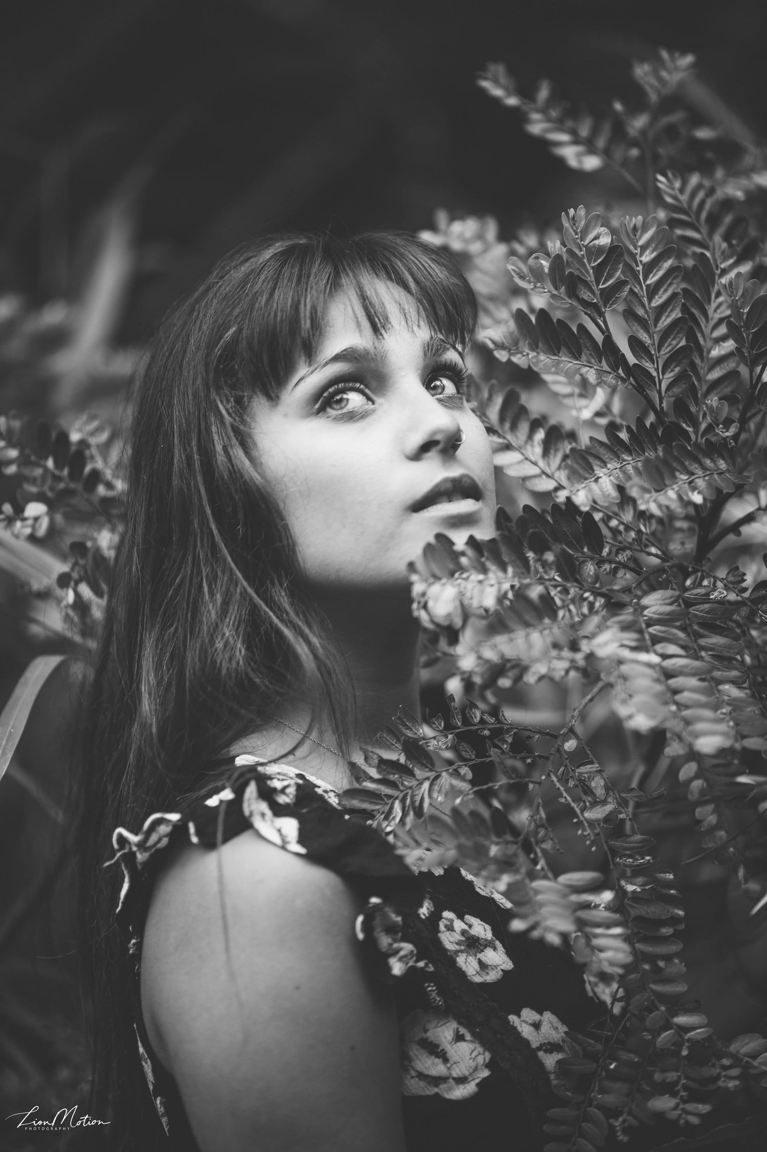 Black and white portrait of a young woman gazing upward surrounded by leafy plants.