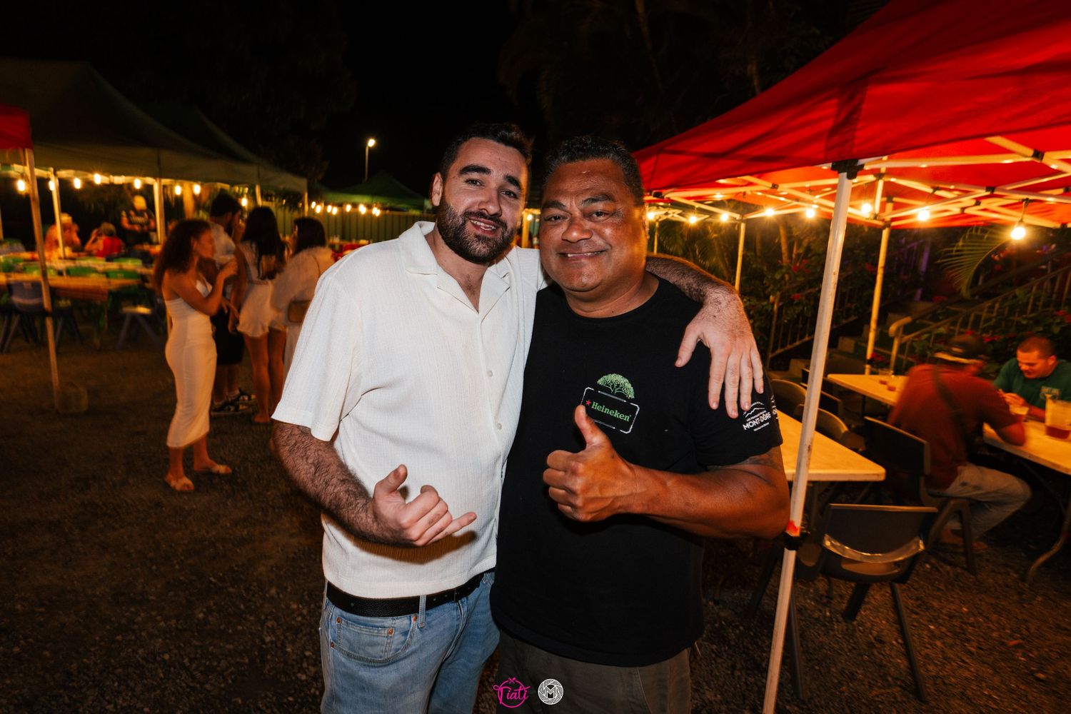 Two men smiling and posing together with thumbs up at an outdoor evening gathering.