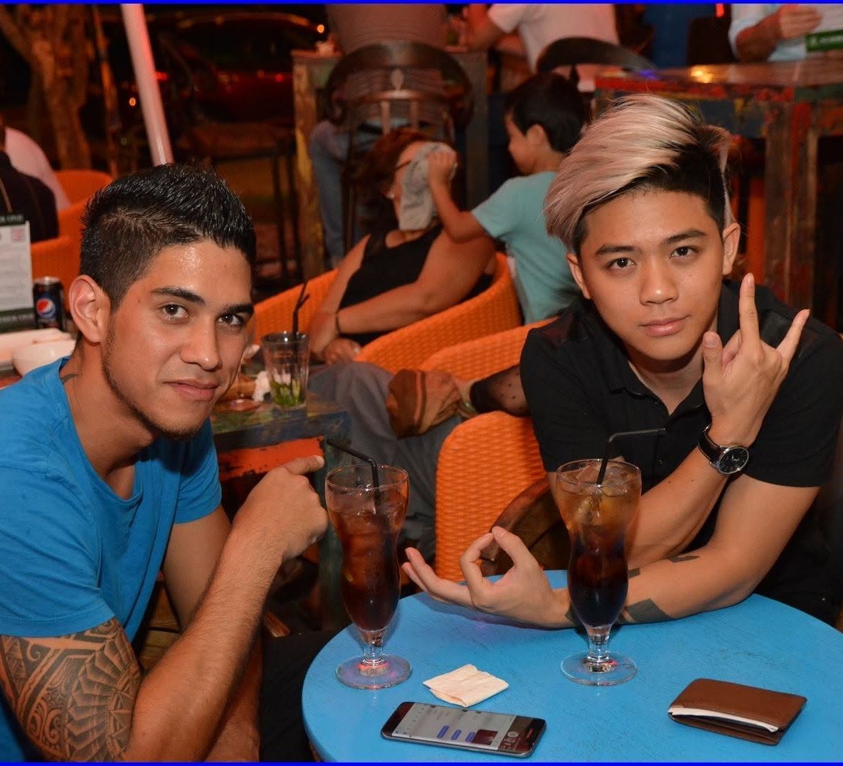 Two young men smiling at camera while sitting at blue table with tropical drinks.