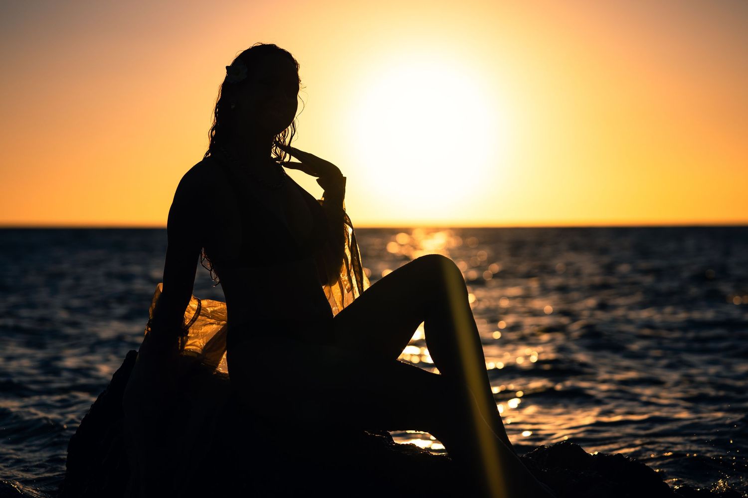 Silhouette of woman sitting on rocks with surfboard during golden ocean sunset.
