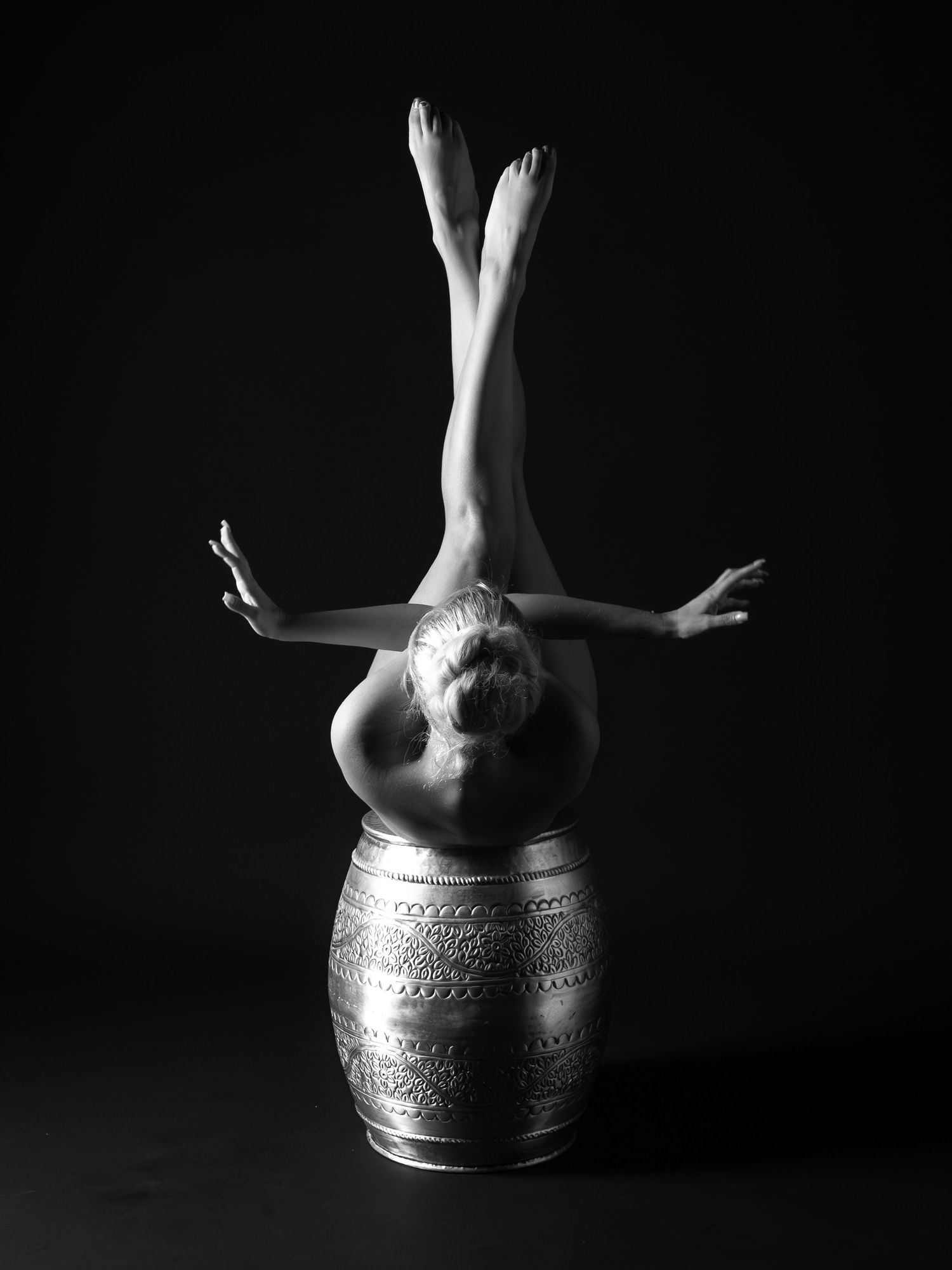Black and white artistic photo of nude woman balancing inverted on ornate barrel.