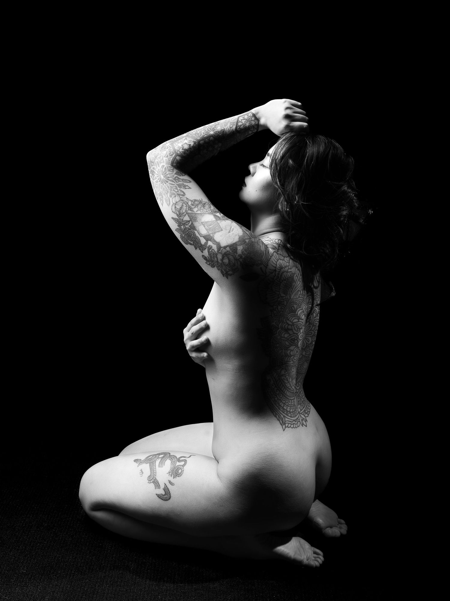 Black and white artistic nude photo of a tattooed woman sitting gracefully with arms raised against dark background.