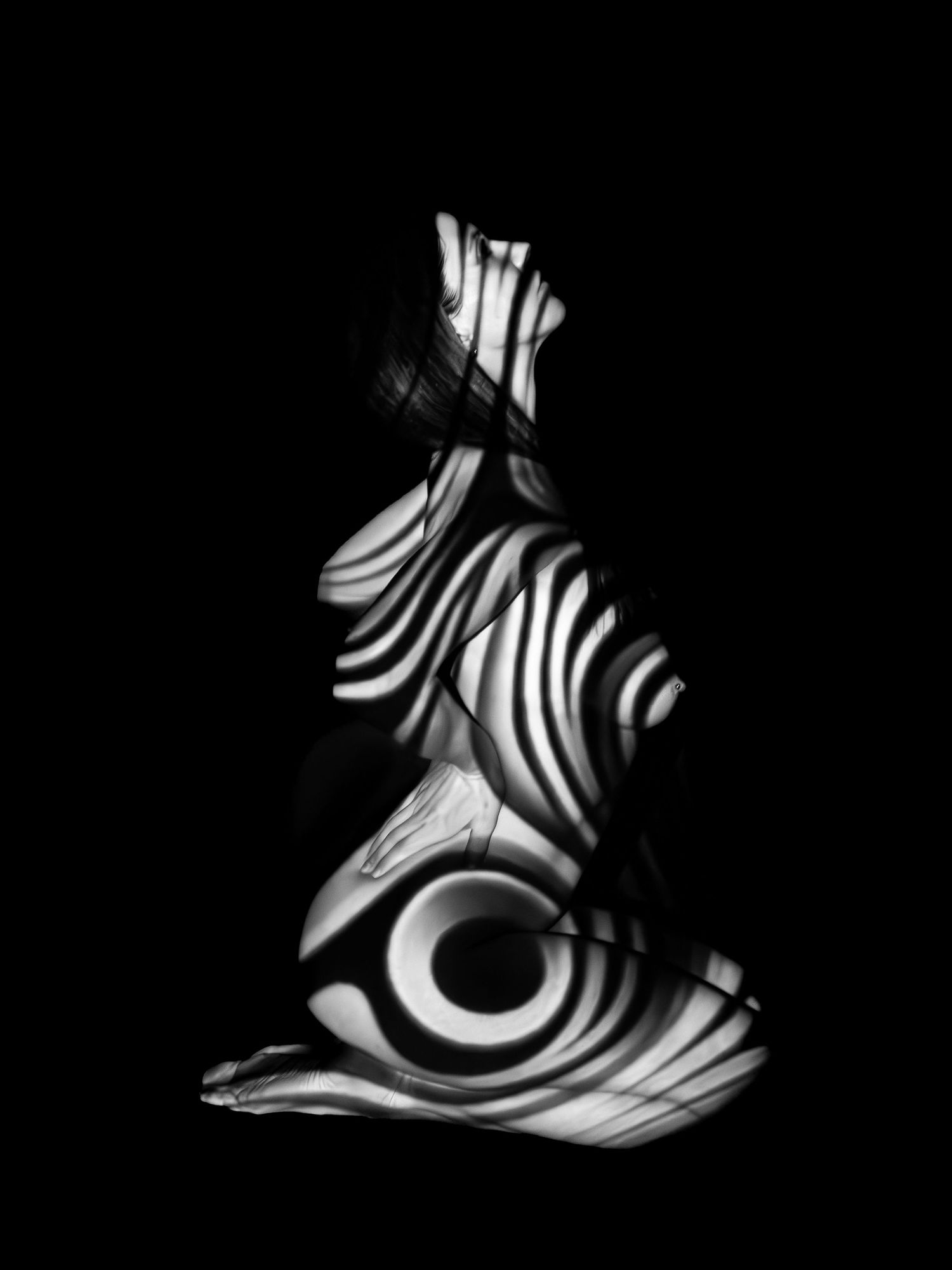 Black and white abstract fine art nude with bold zebra stripe light projections on dark background.
