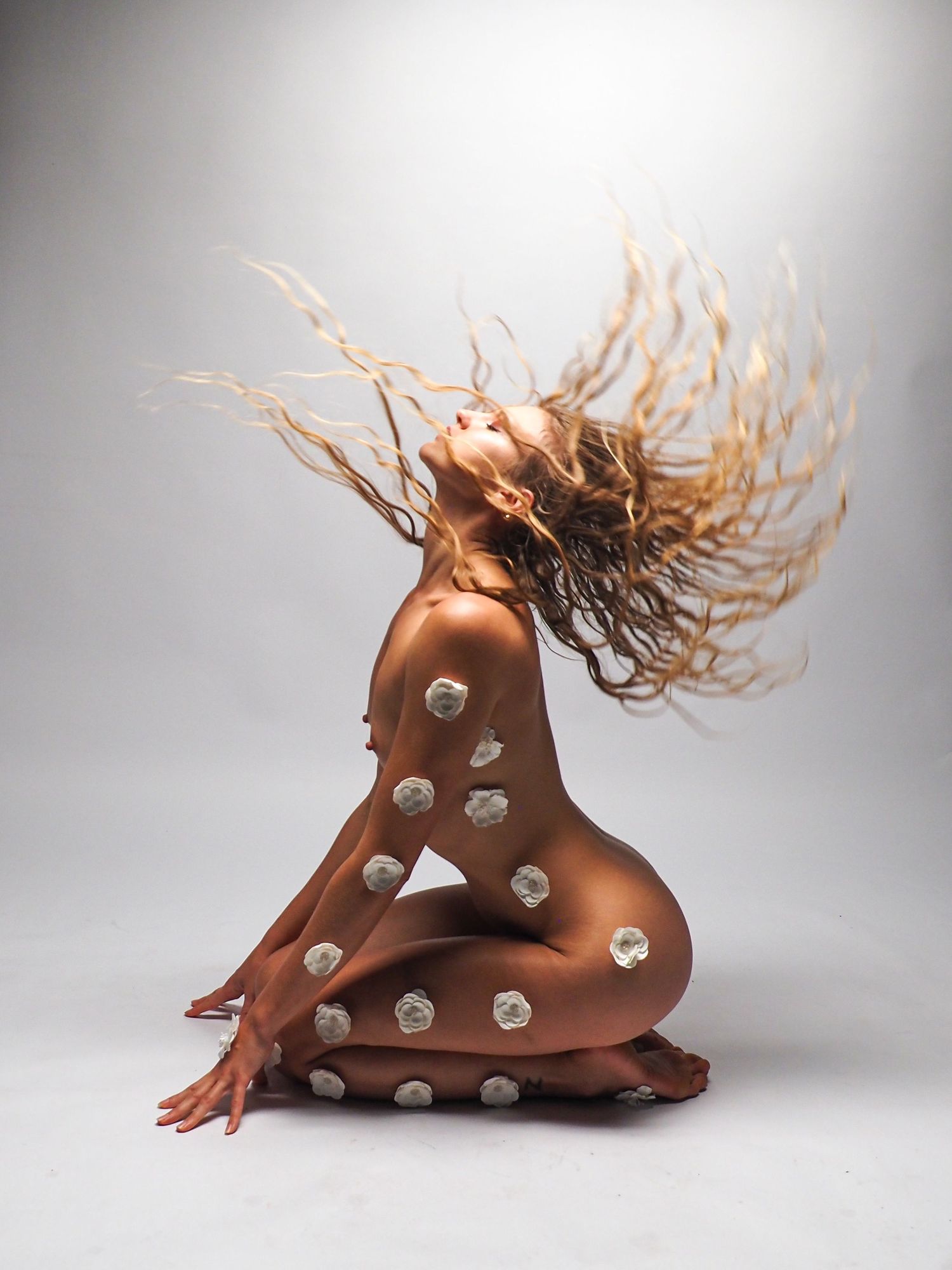 Nude woman with copper body paint and white roses tossing long hair dramatically.