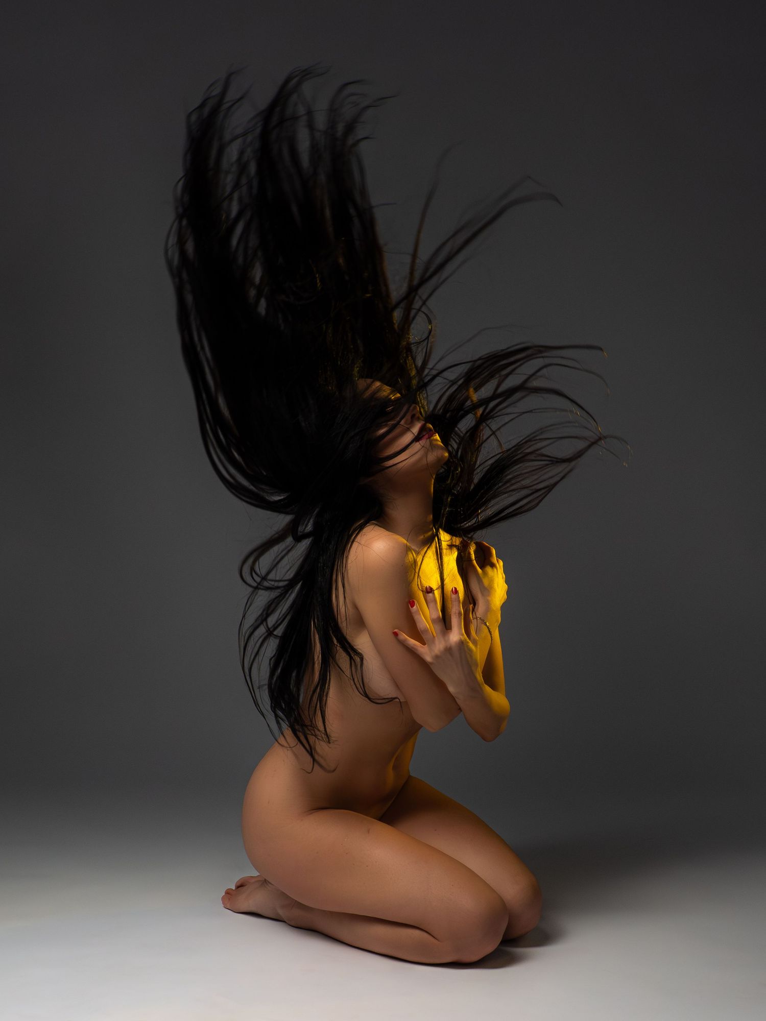 Fine art nude with long black hair dramatically flipped upward holding yellow fabric on gray background.