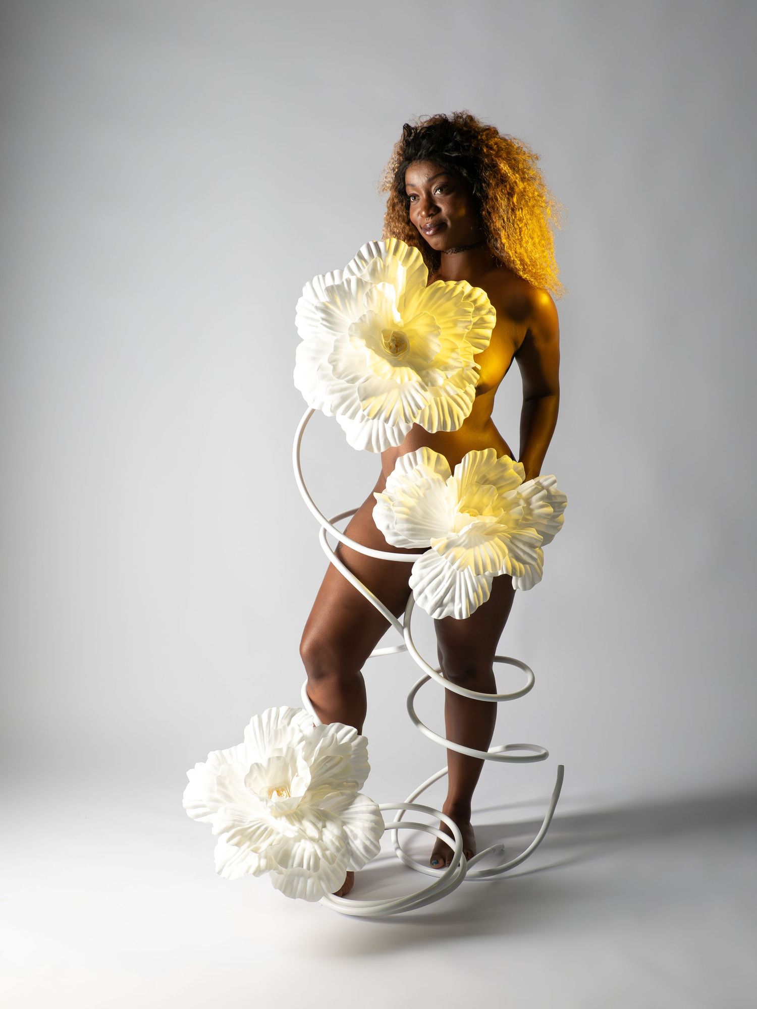 Woman with yellow body paint poses with large white flowers on spiral stand.