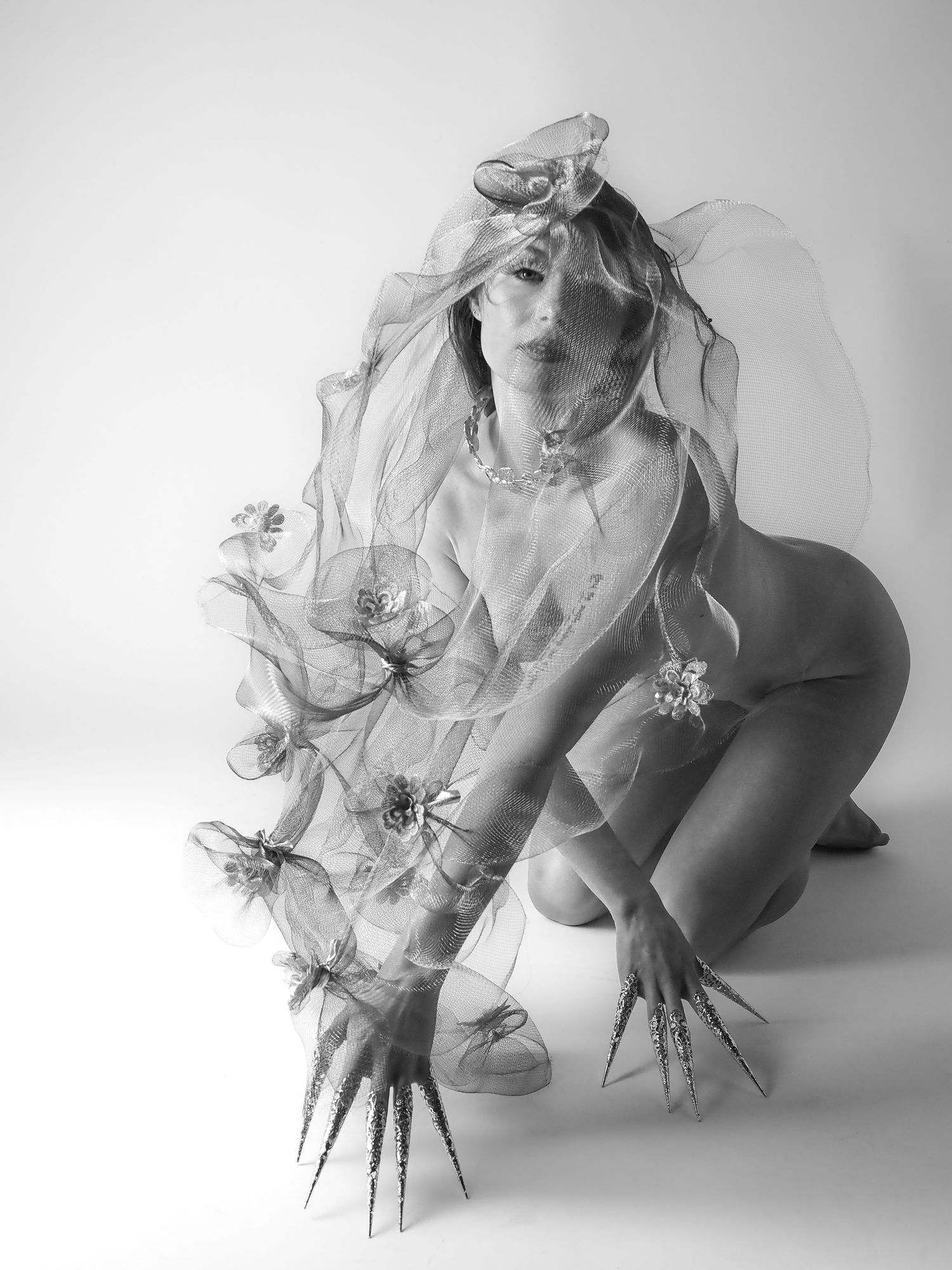 Black and white artistic nude with sheer floral veil and spiked accessories in studio.