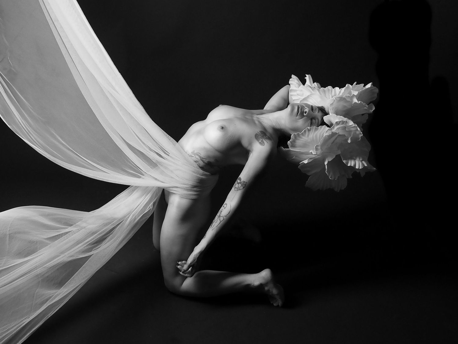 Black and white fine art nude with flowing white fabric and floral headpiece in dramatic pose.