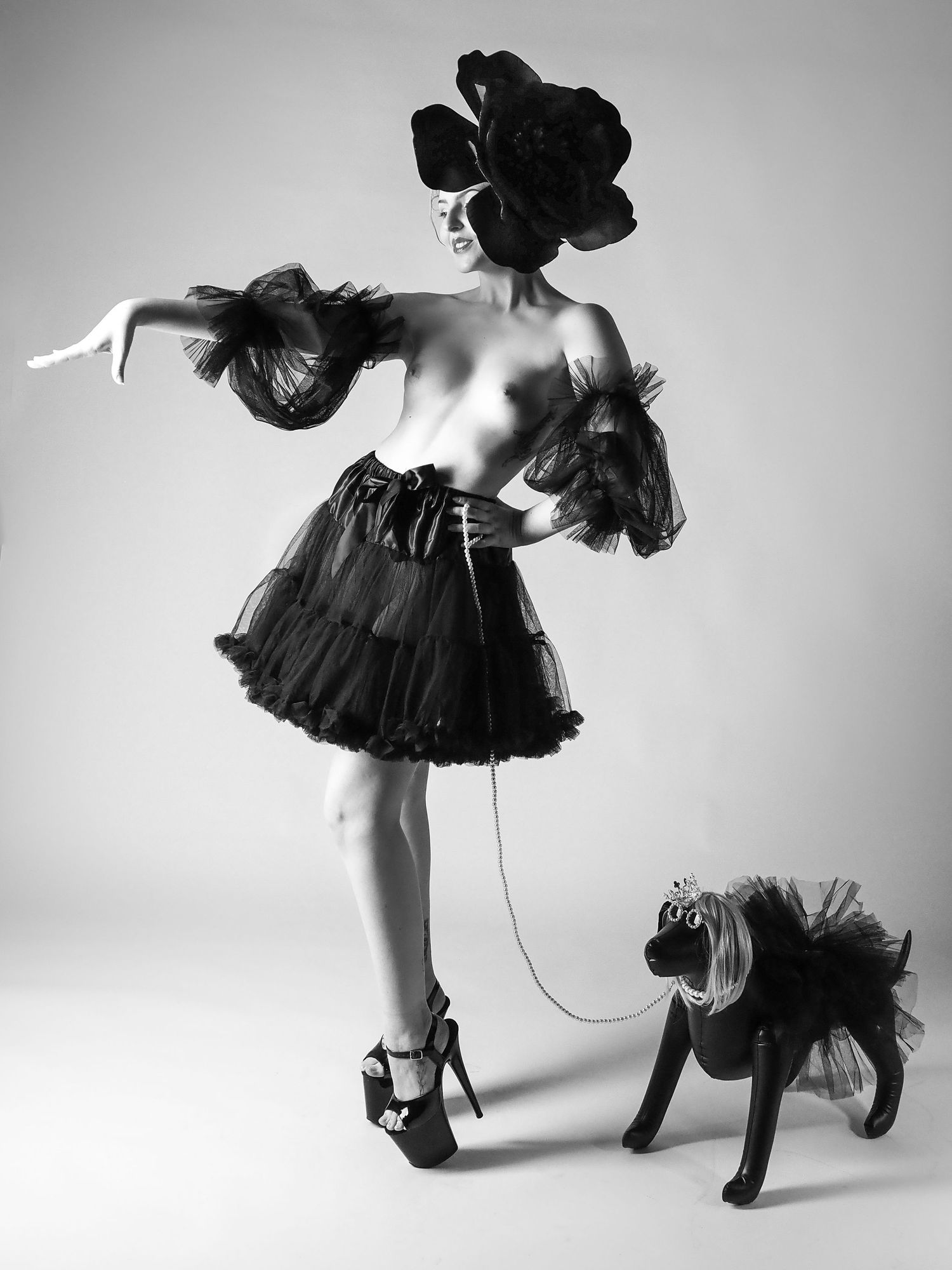 Black and white fashion photo of woman in tulle skirt and floral headpiece posing with decorative dog on leash.