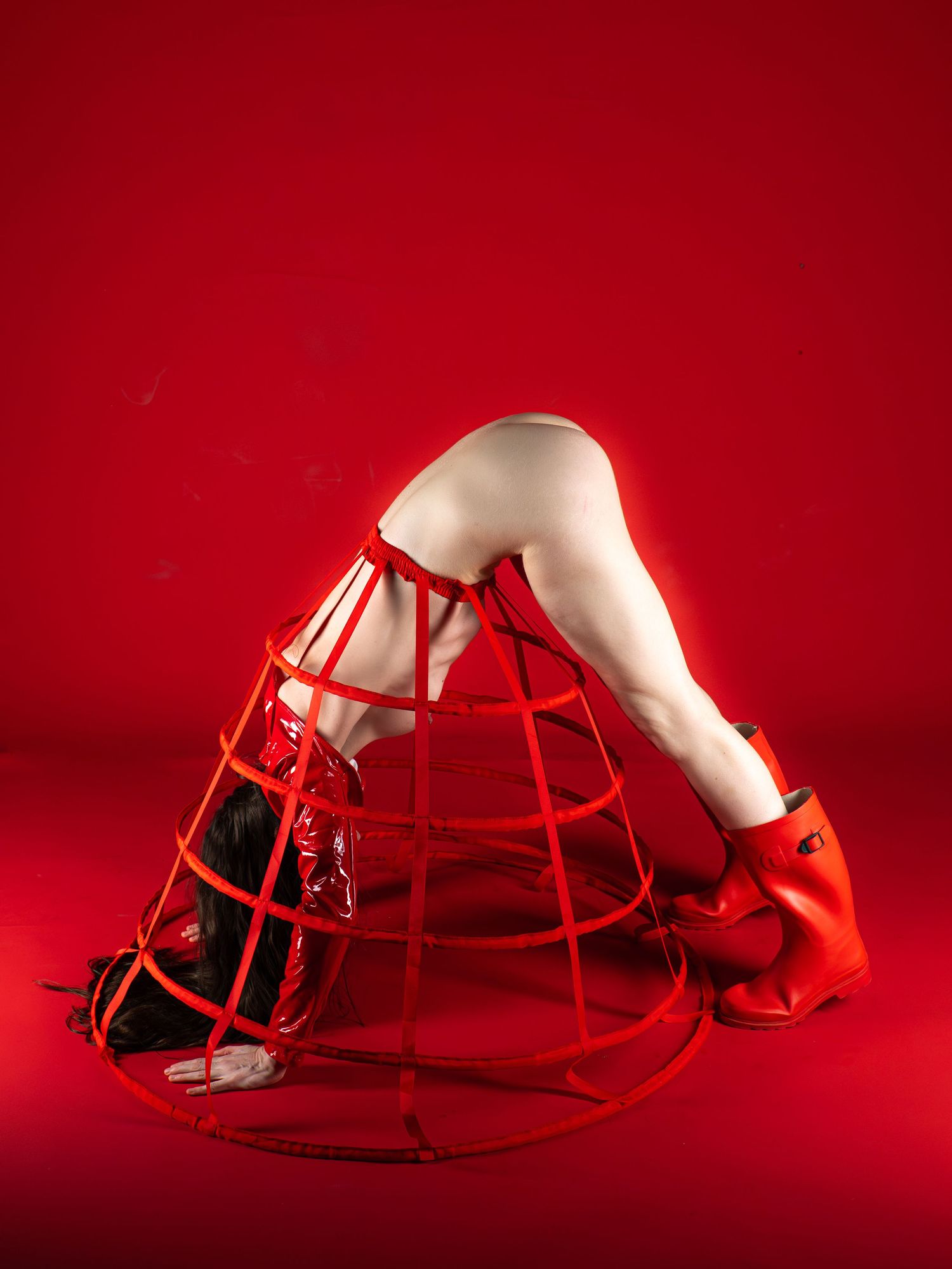 Fine art photo of legs emerging from a red hoop crinoline cage structure on a red background.