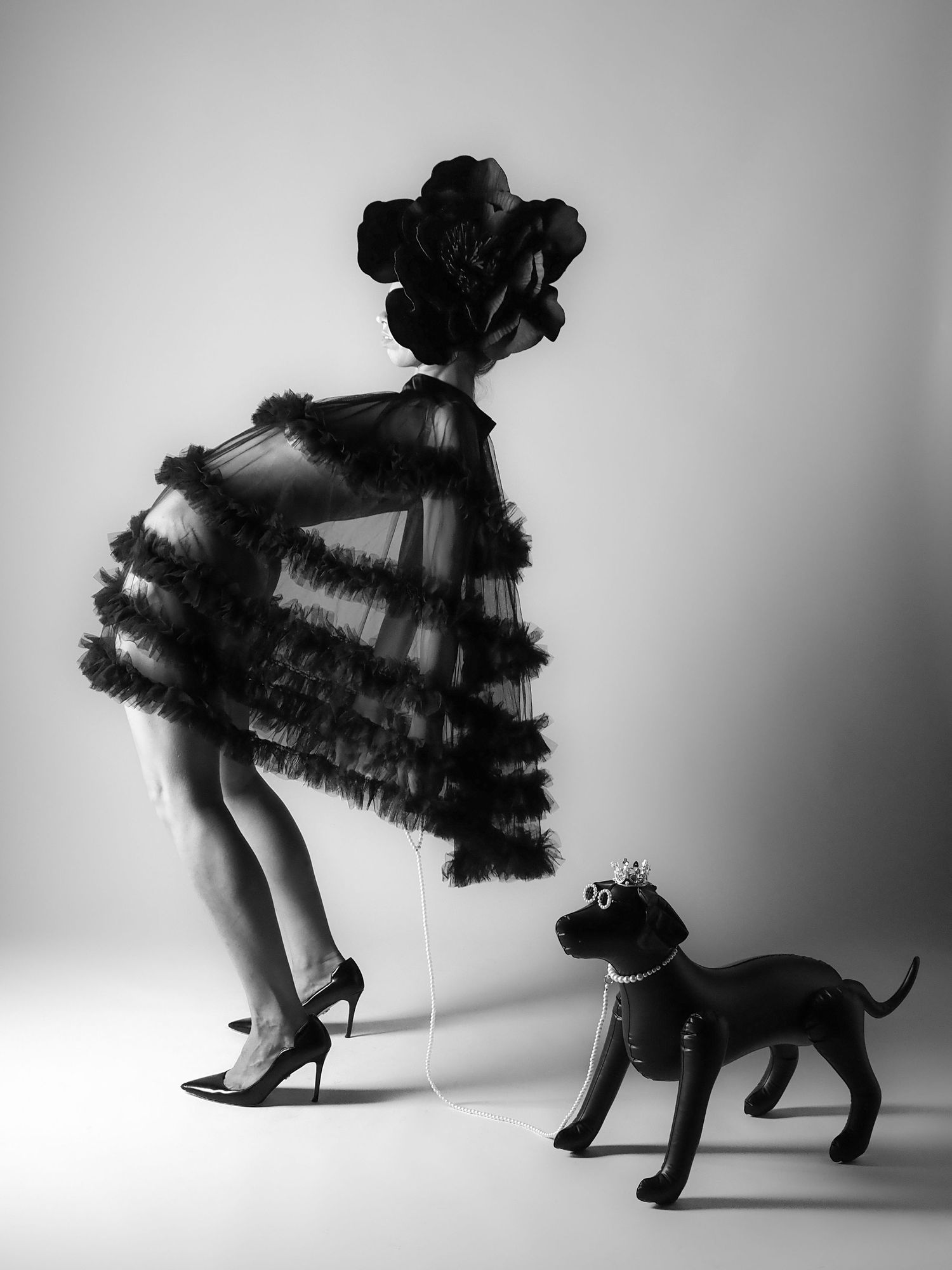 Black and white photo of figure in ruffled tulle dress with floral headpiece walking a toy dog on a string.