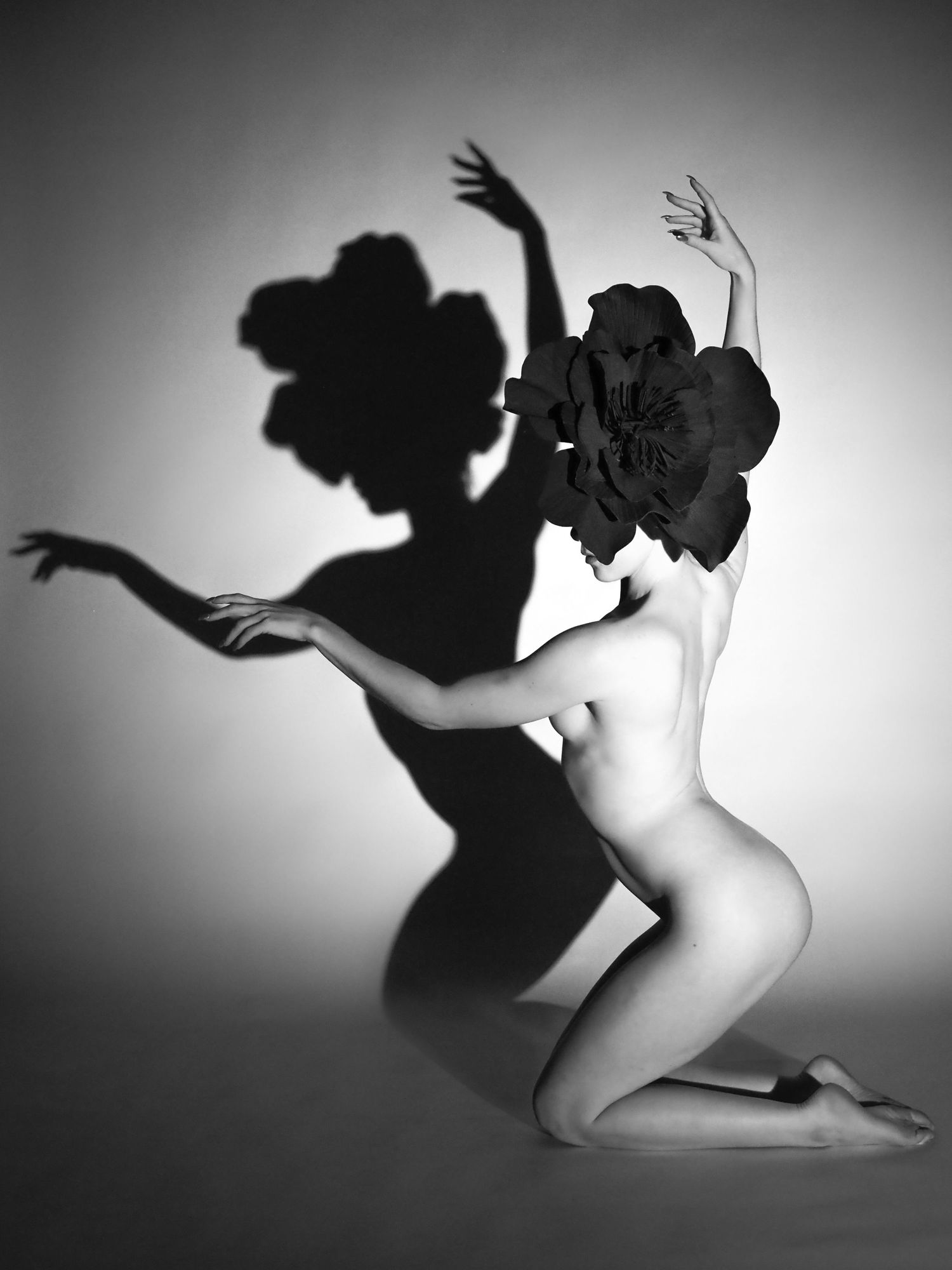 Black and white artistic nude with large floral headpiece casting dramatic shadow on studio wall.