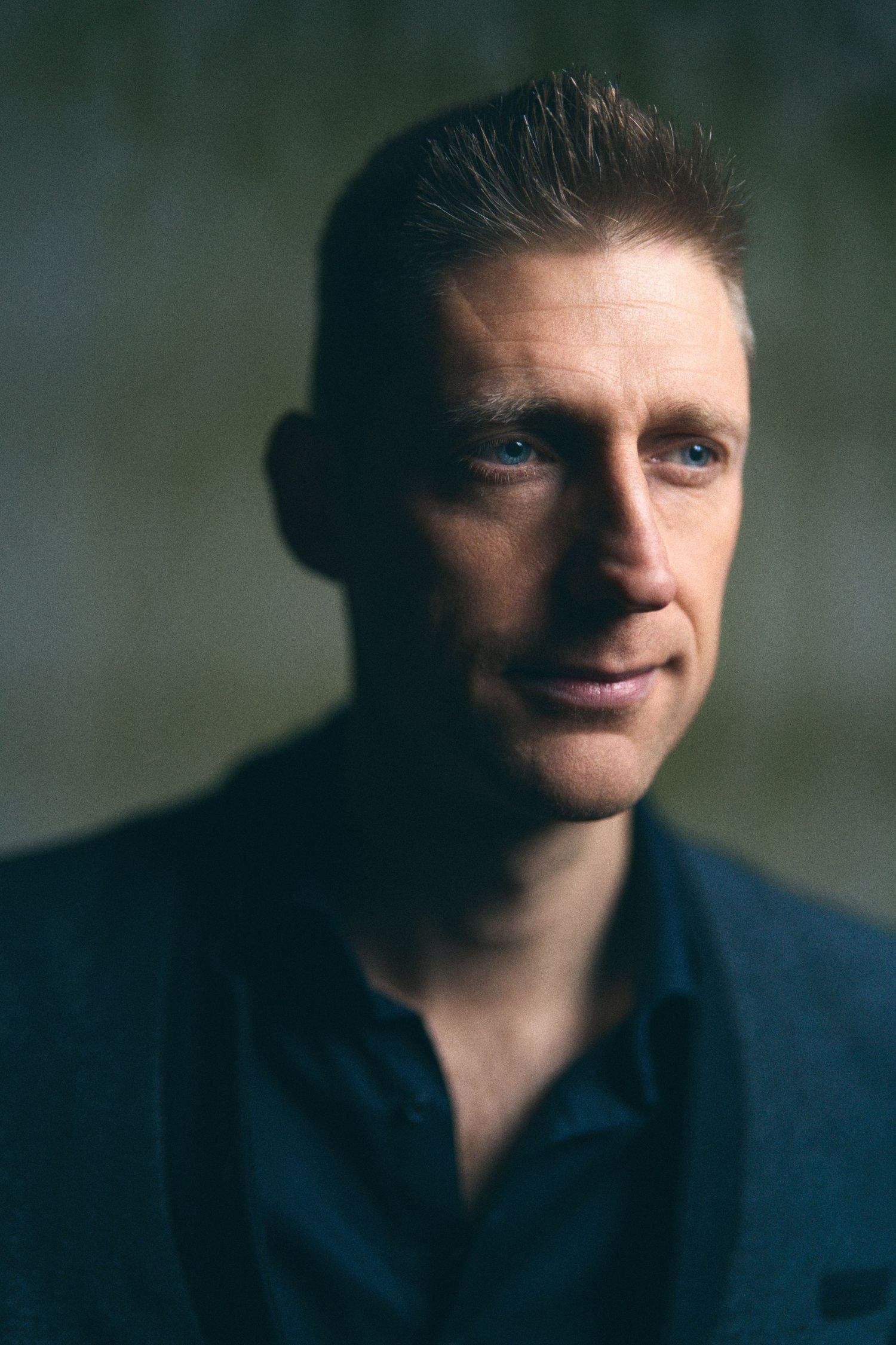 Blue-eyed man in dark shirt with slicked-back hair posing for moody portrait against textured wall.