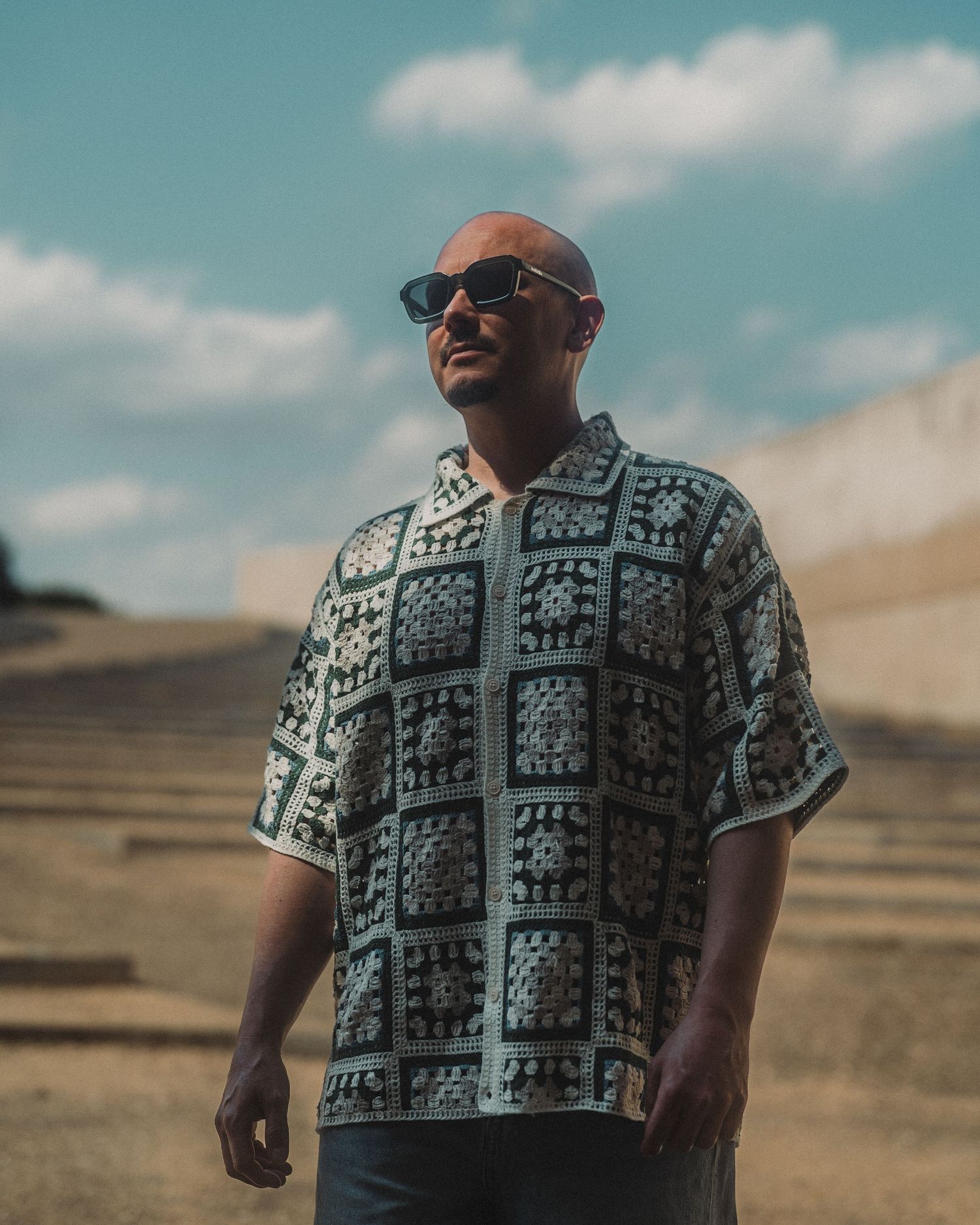 Bald man wearing sunglasses and a patterned crochet shirt standing outdoors on sunny day.