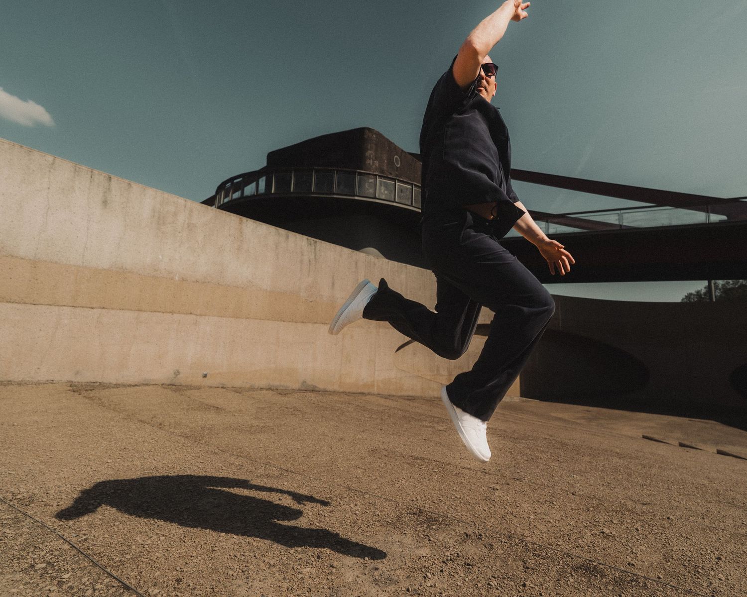 Person jumping mid-air near concrete wall and bridge in sunlight.