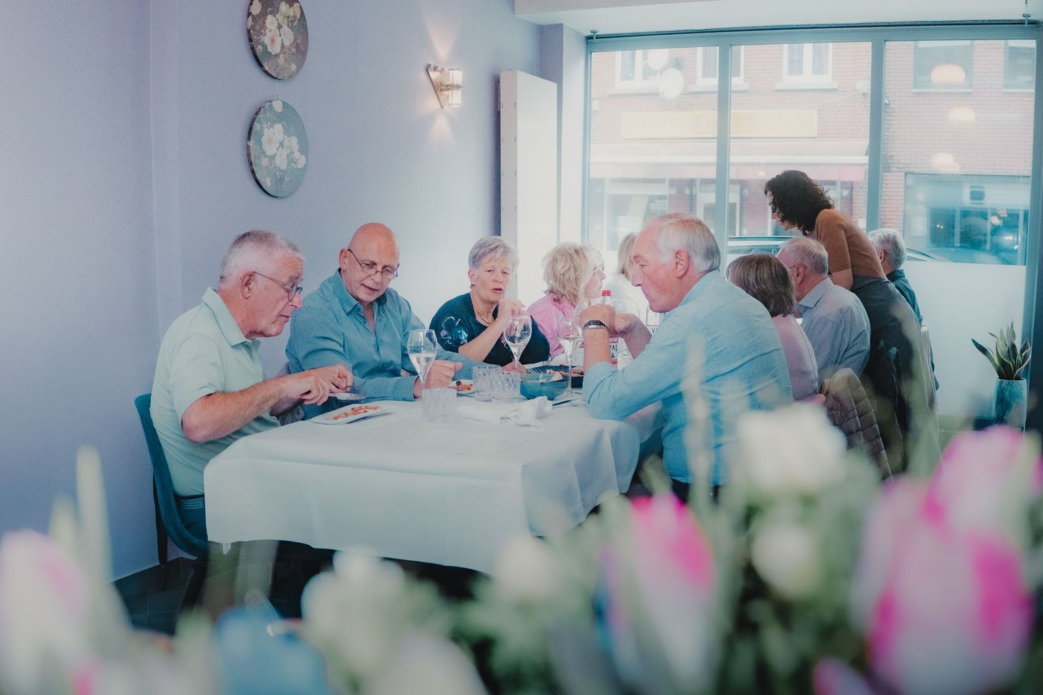 Elderly group enjoying a social gathering at a white table in a bright dining room with tulips in the foreground.