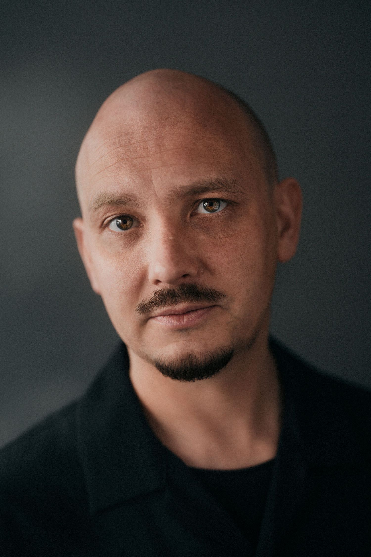 Bald man with goatee in black jacket posing for dark portrait.